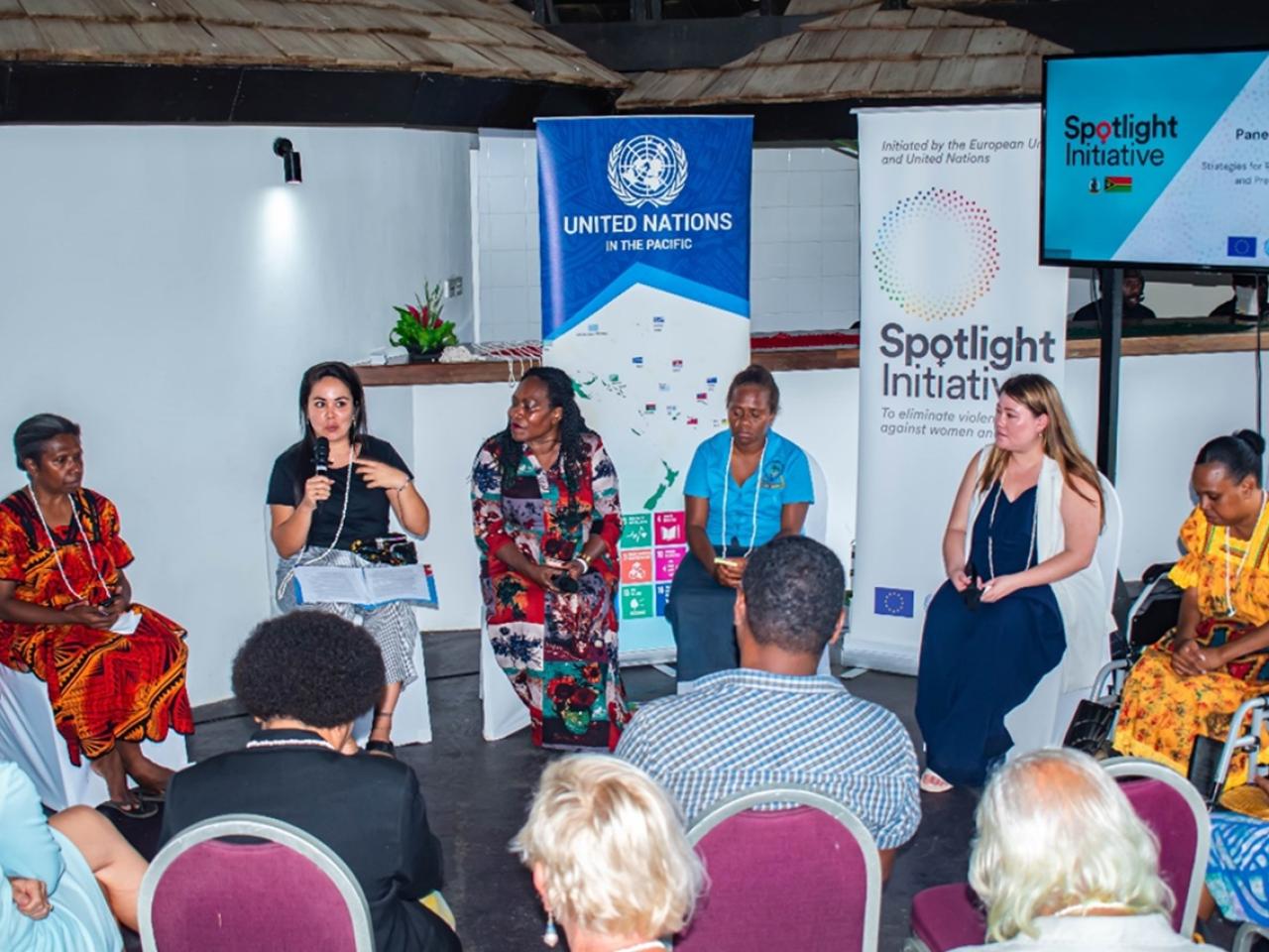 Six women panels sitting in front of audience, one of them, the moderator speaking with a microphone.