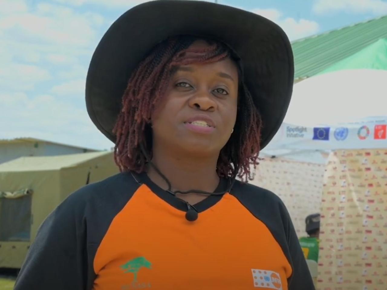 Woman in black hat and orange and black t-shirt speaking into camera