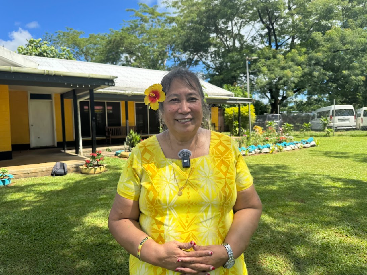 Lina Chang smiles at camera. She wears a yellow dress and flower behind her ear and stands in front of a yellow building.