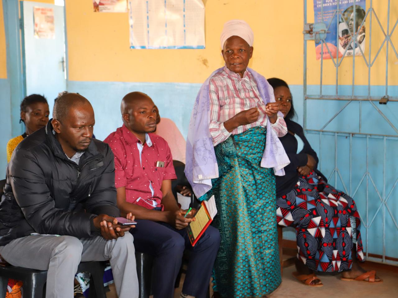 A woman stands and speaks next to two seated men.
