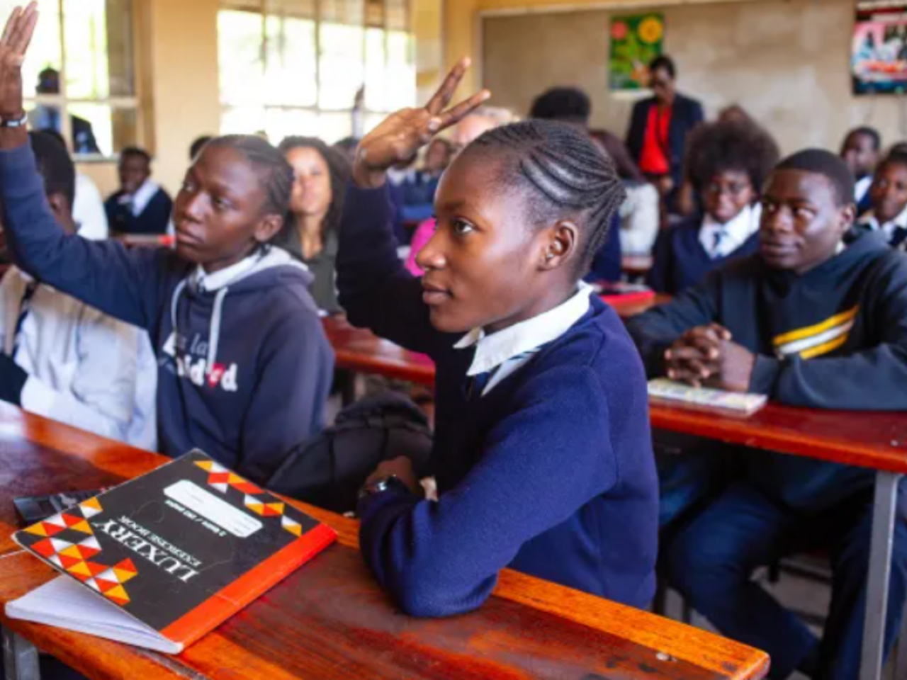 Girls in unform sitting behind desks in classroom with hand raised 