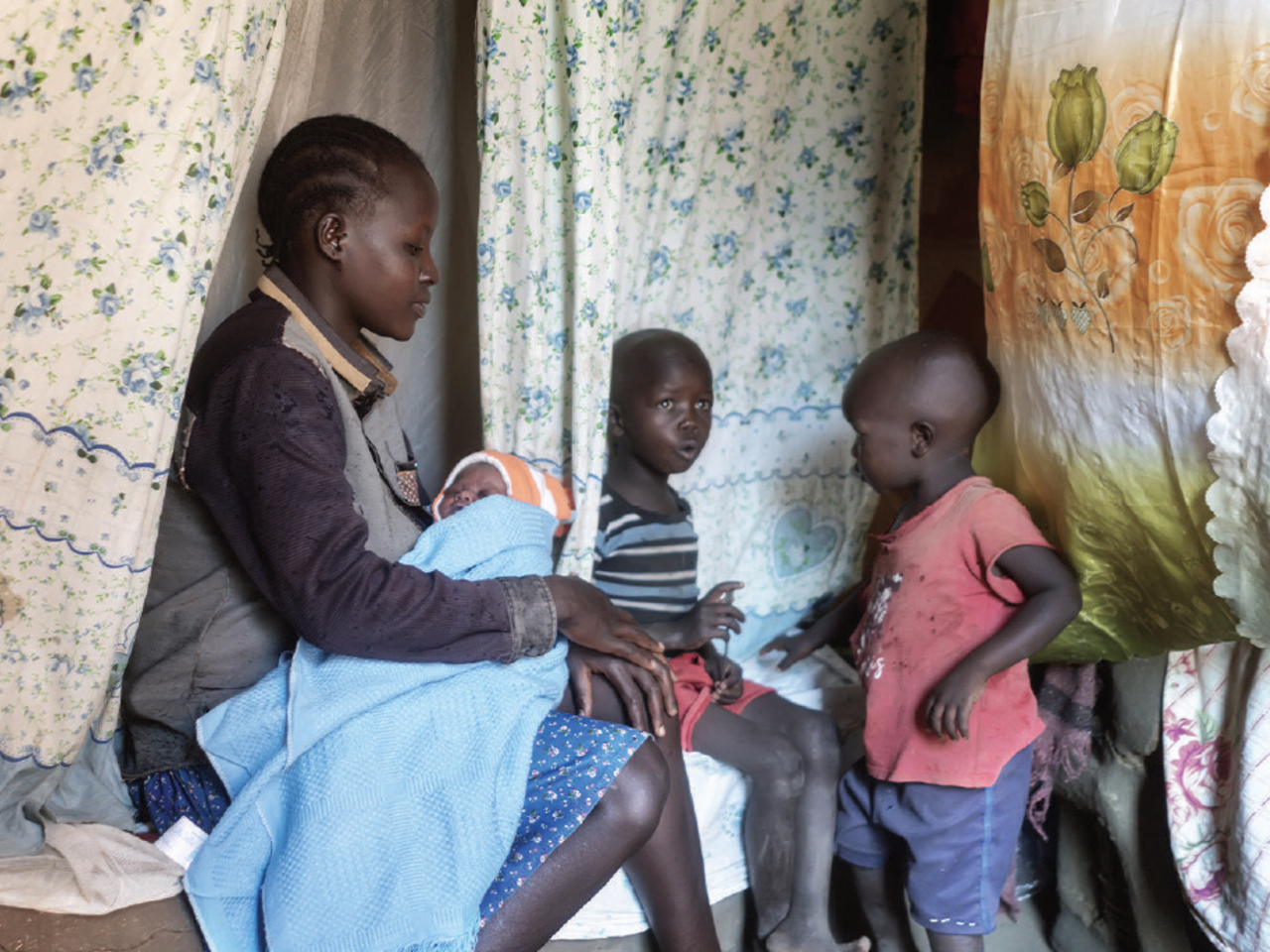 Alinda is seated and holds a baby in her arms with two children beside her