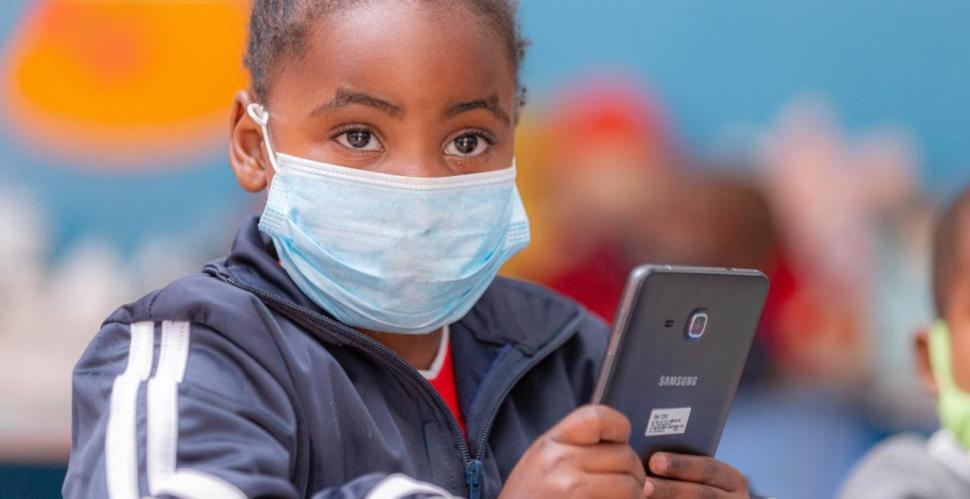 A young child wearing a surgical mask holds a phone