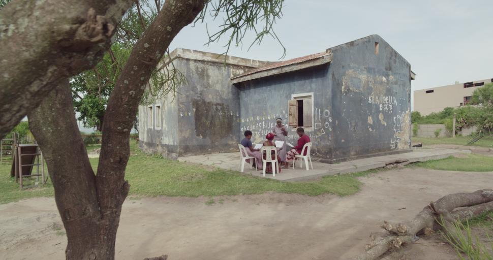 womne seated outside a small building with a tree in the foreground