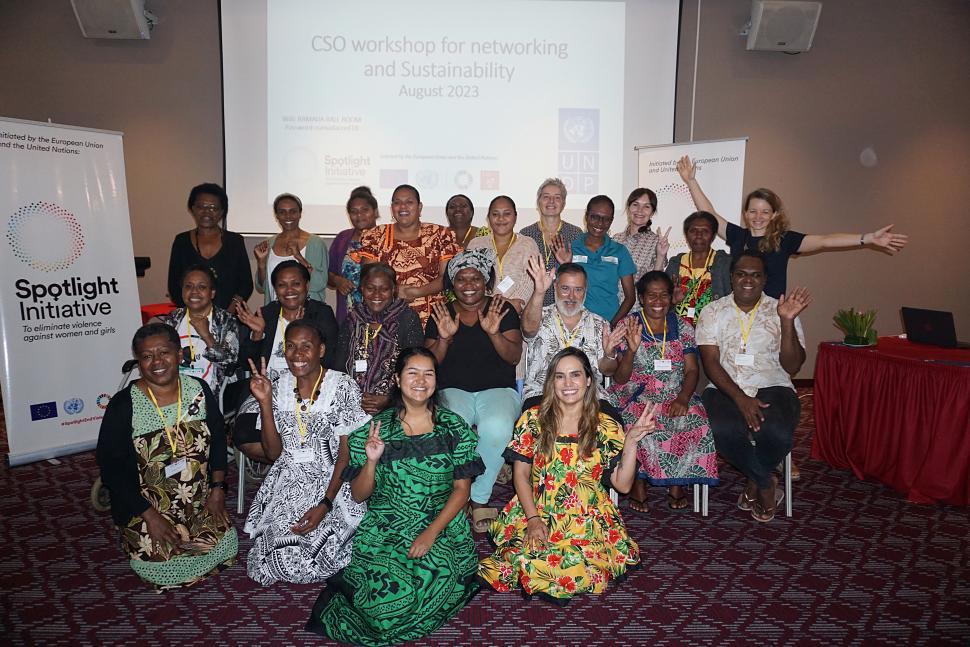 A group photo of a workshop, some sitting on the floor, the other standing, with a Spotlight Initiative pull-up banner on the left back and a bright screen on the wall of the room behind the group.