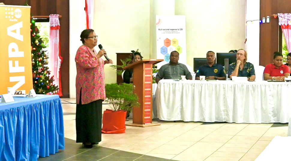 A woman standing in front of audience speaking with microphone and three men and a woman at the side table in a room for the ceremony. 