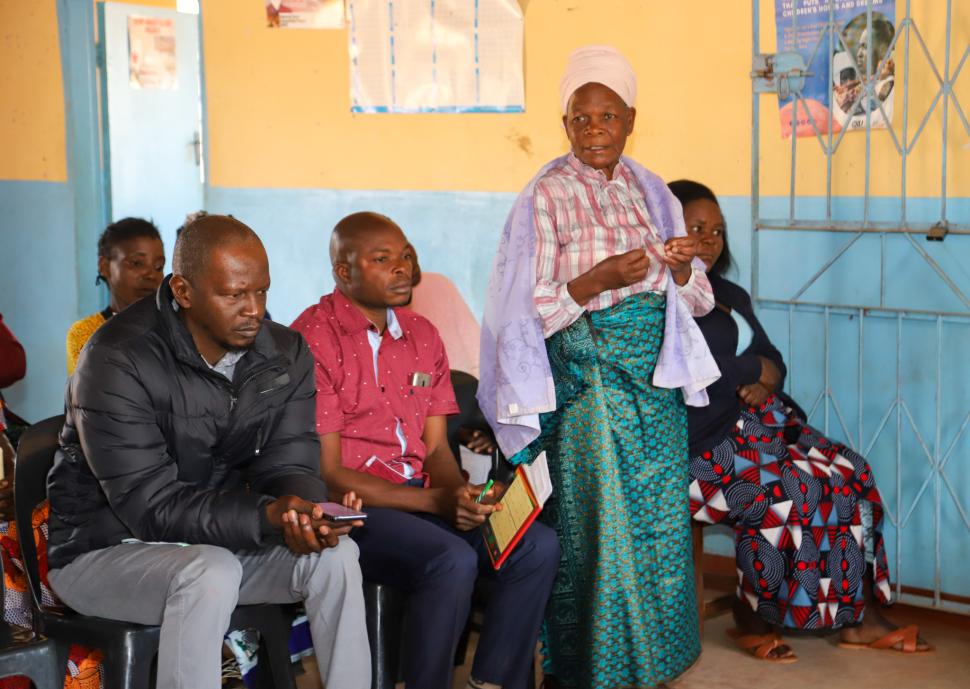 A woman stands and speaks next to two seated men.