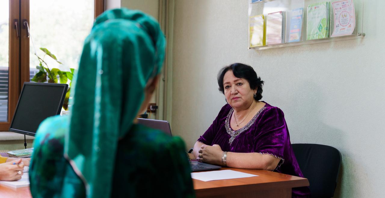 A woman in a purple dress behind a desk speaking to a woman with a green head covering