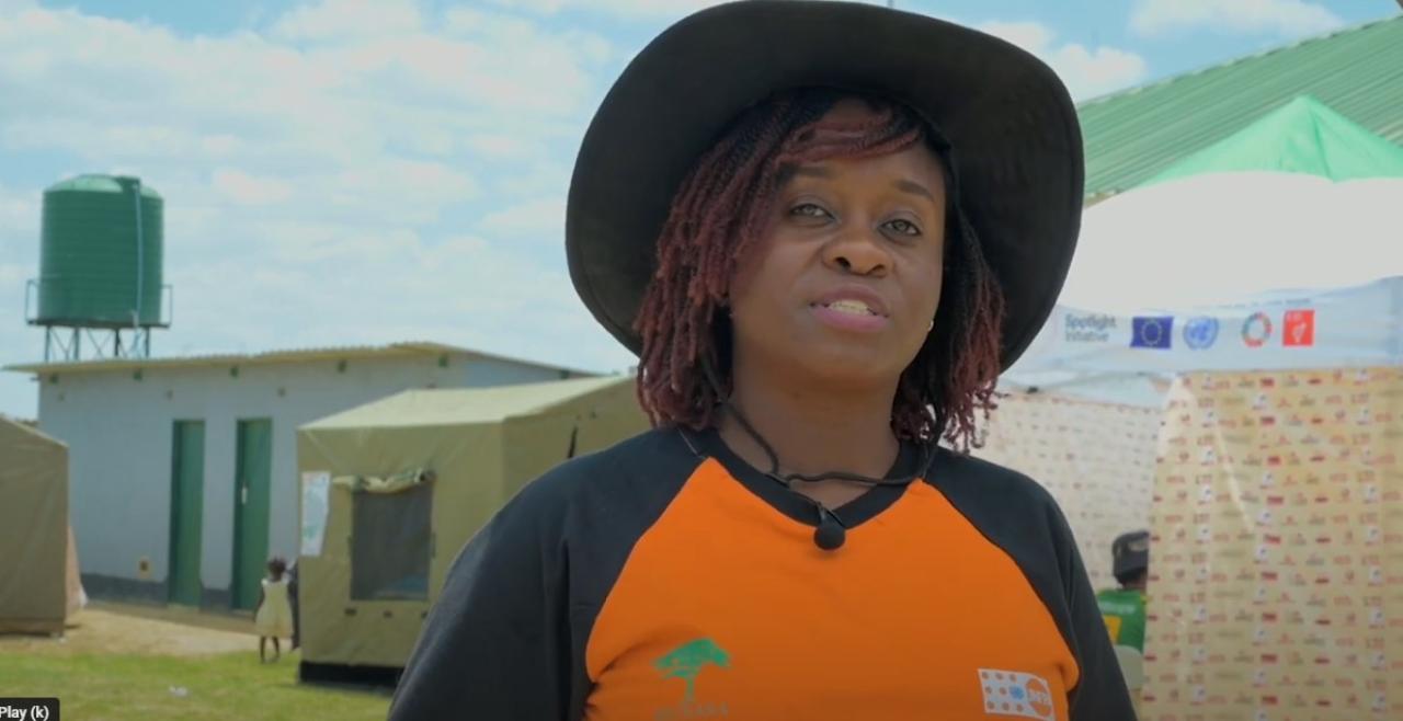 Woman in black hat and orange and black t-shirt speaking into camera