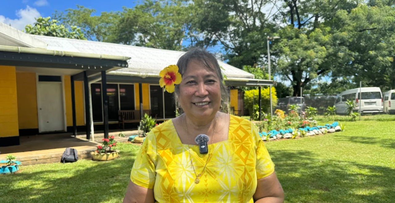 Lina Chang smiles at camera. She wears a yellow dress and flower behind her ear and stands in front of a yellow building.