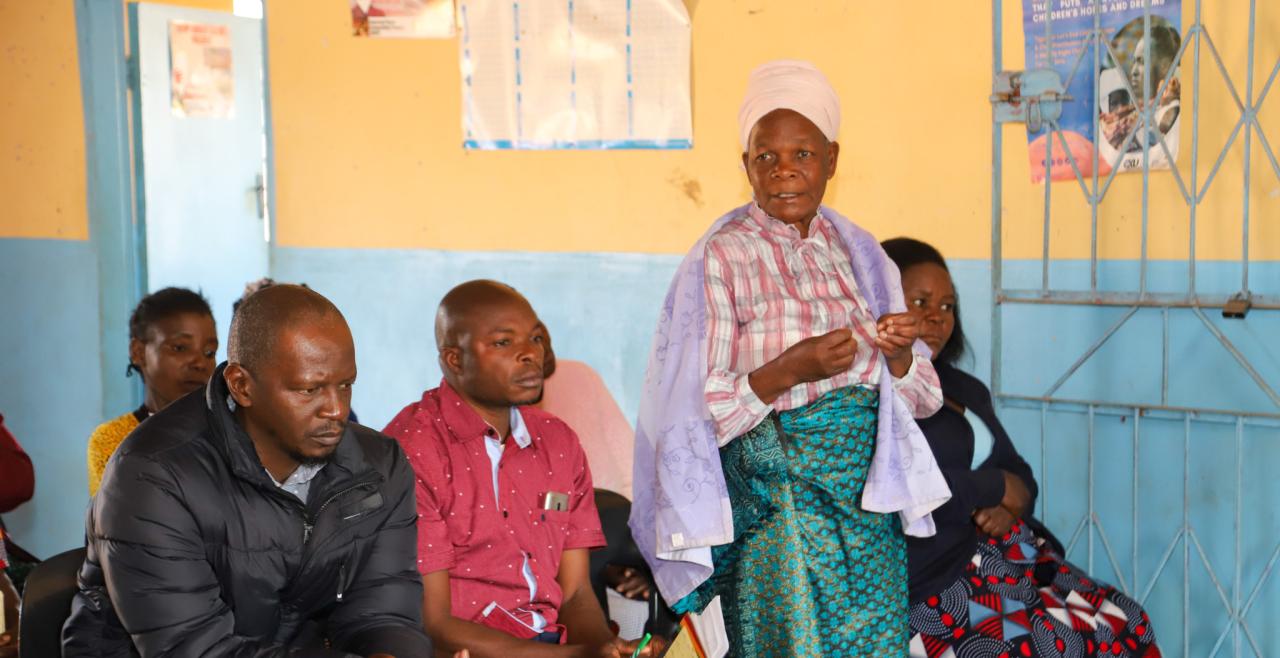 A woman stands and speaks next to two seated men.