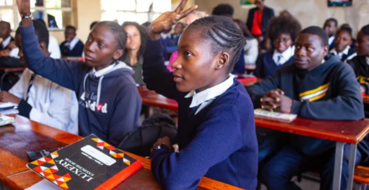 Girls in unform sitting behind desks in classroom with hand raised 