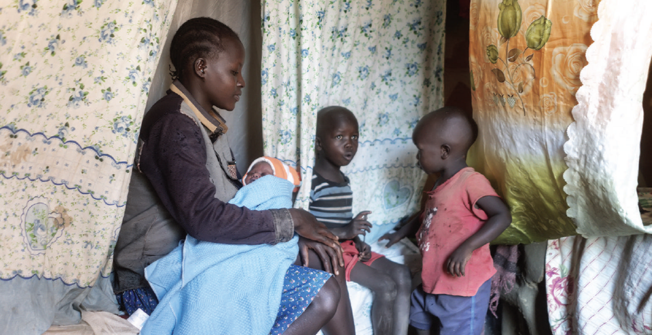 Alinda is seated and holds a baby in her arms with two children beside her